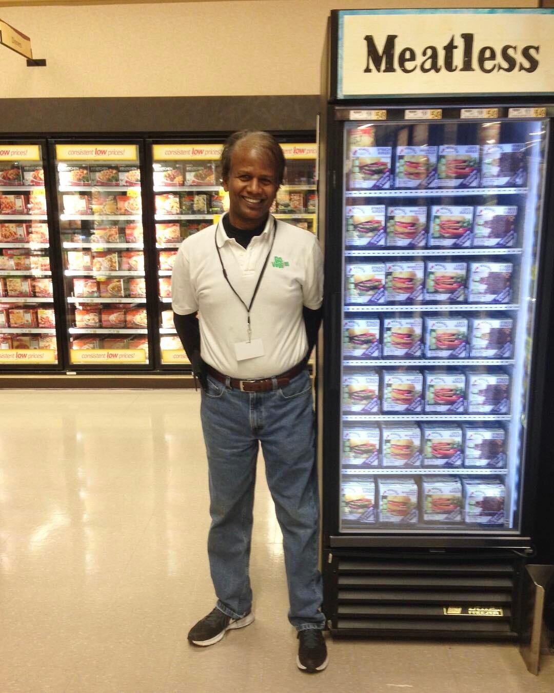 A smiling man stands beside a freezer labeled "Meatless," showcasing a variety of meatless food options in a grocery store aisle. The background features additional freezers filled with frozen meals.